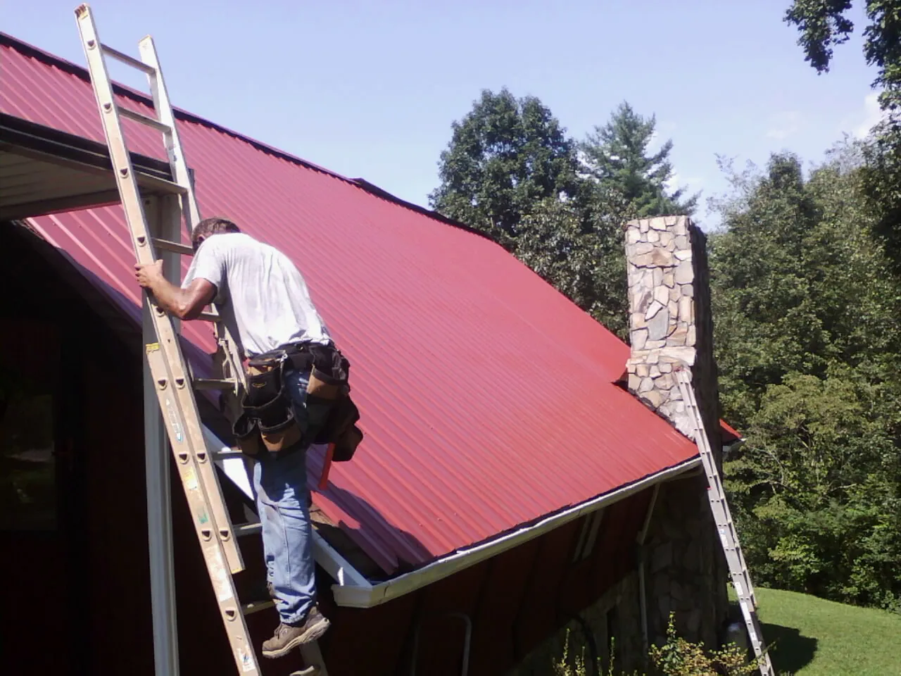 Roofer on a ladder beside a red metal roof and stone chimney — Soffit & Fascia Repair by Roofing Brothers in Manhattan
